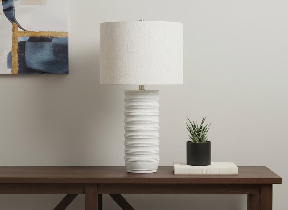 28" Cream Cylinder Table Lamp With Cream Drum Shade displayed on a dark wood tabletop in a furnished interior against an off-white wall.