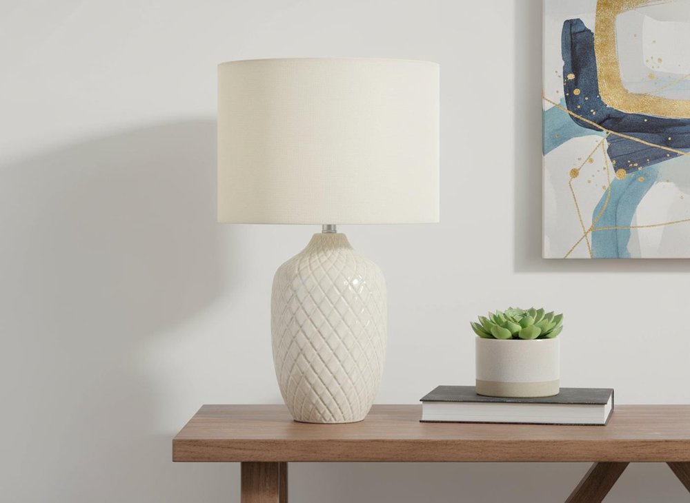 25" Cream Ceramic Urn Table Lamp With Cream Drum Shade displayed on a tabletop in a furnished interior against an off-white wall.