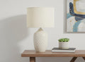 25" Cream Ceramic Urn Table Lamp With Cream Drum Shade displayed on a tabletop in a furnished interior against an off-white wall.