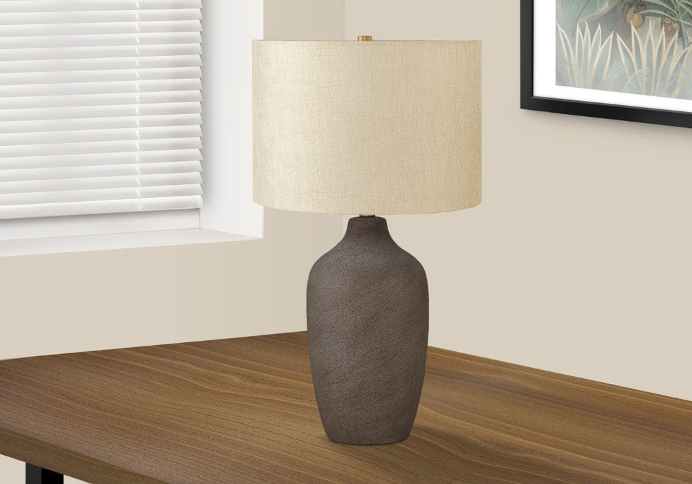 27" Gray Ceramic Urn Table Lamp With Beige Drum Shade displayed on a wood tabletop in a furnished interior against a cream wall.