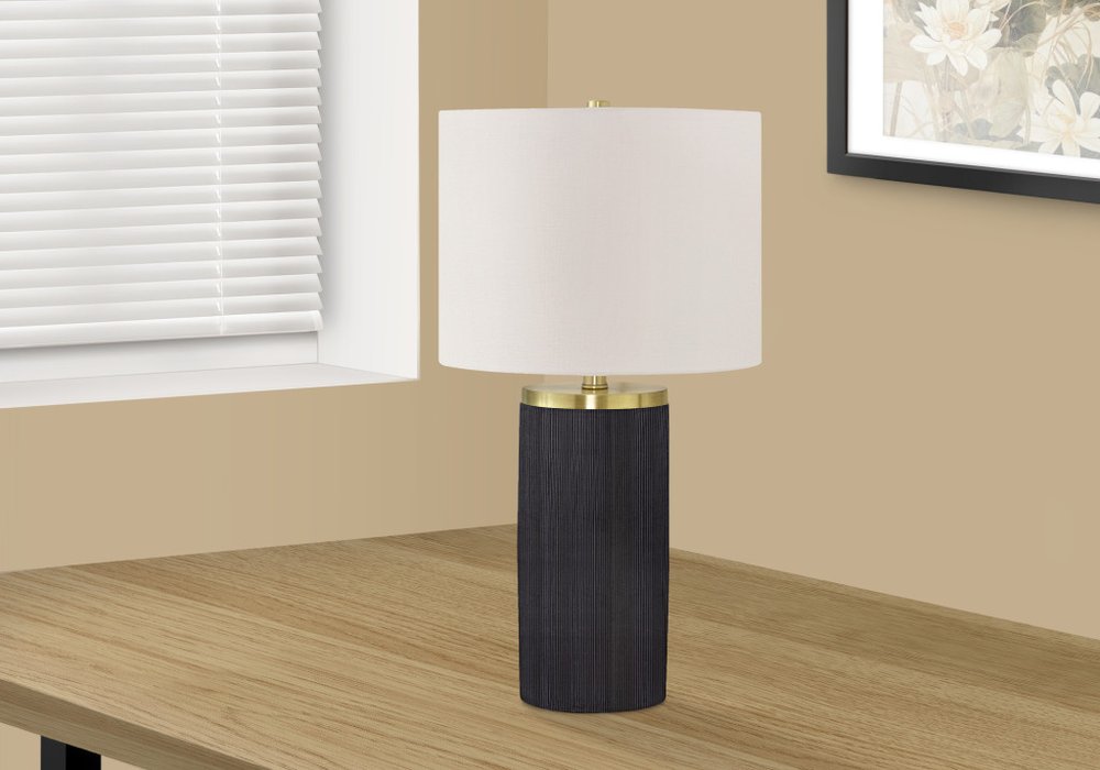 24" Black and Gold Concrete Cylinder Table Lamp With Ivory Drum Shade displayed on a wood tabletop in a furnished interior against an off-white wall.