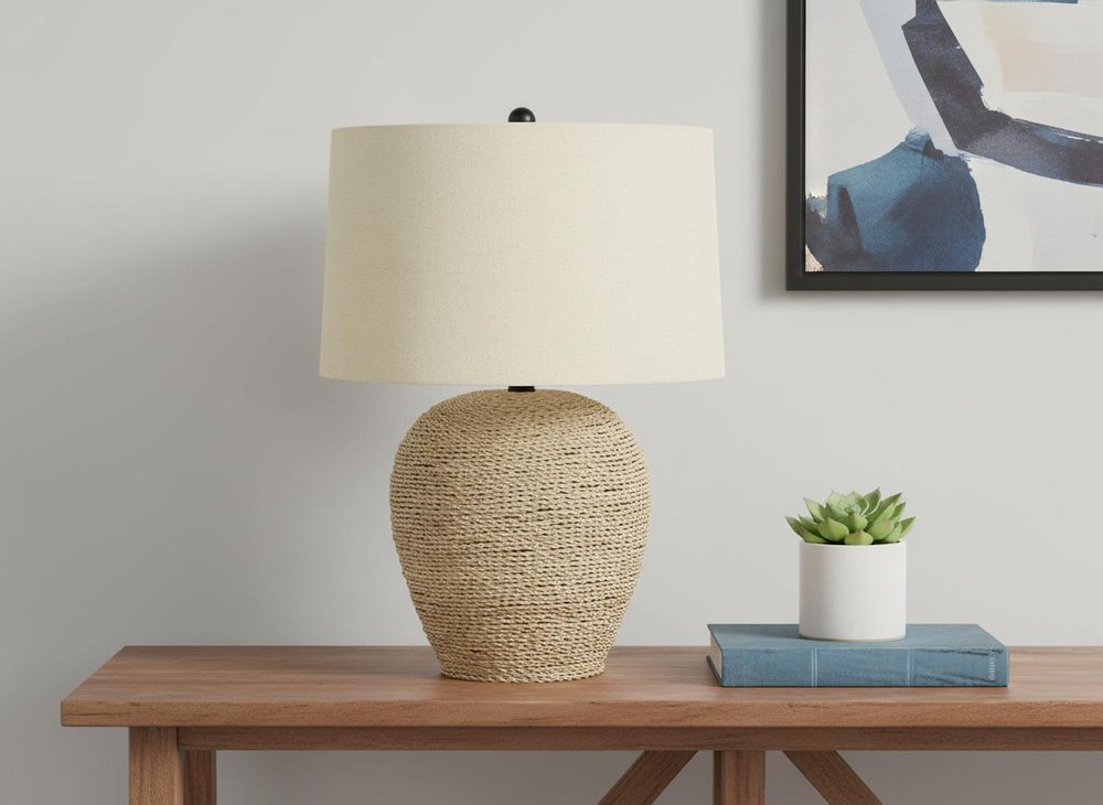25" Beige Rattan Round Table Lamp With Beige Drum Shade displayed on a tabletop in a furnished interior against an off-white wall.
