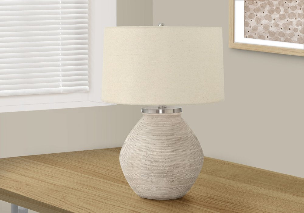 25" Cream Concrete Round Table Lamp With Beige Drum Shade displayed on a wood tabletop in a furnished interior against an off-white wall.