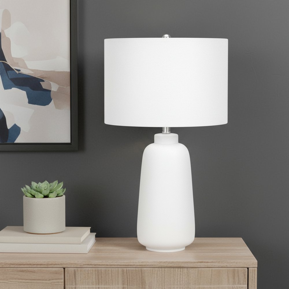 26" Cream Ceramic Cylinder Table Lamp With Cream Drum Shade displayed on a wood tabletop in a furnished interior against a gray wall.
