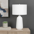 26" Cream Ceramic Cylinder Table Lamp With Cream Drum Shade displayed on a wood tabletop in a furnished interior against a gray wall.