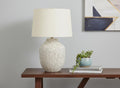 26" Cream Ceramic Round Table Lamp With Cream Empire Shade displayed on a tabletop in a furnished interior against a light gray wall.