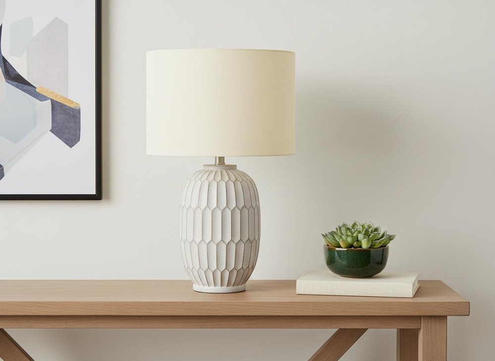 24" Cream Geometric Table Lamp With Cream Drum Shade displayed on a wood tabletop in a furnished interior against an off-white wall.