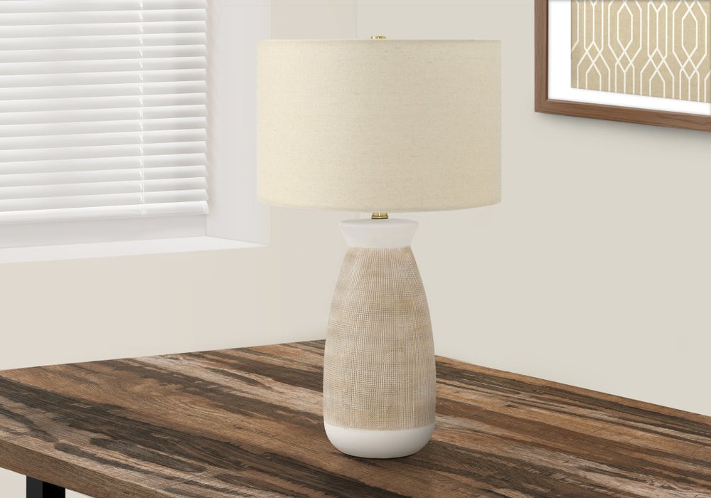 27" Brown and White Ceramic Round Table Lamp With Beige Drum Shade displayed on a dark wood tabletop in a furnished interior against an off-white wall.
