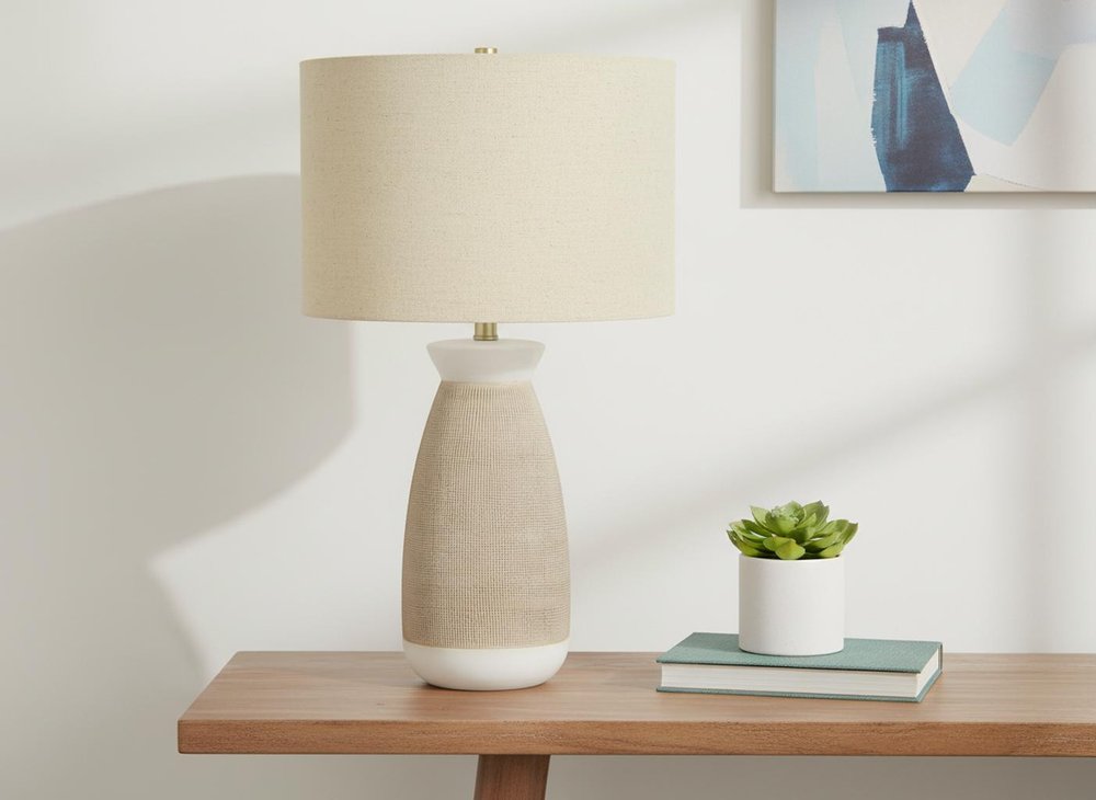 27" Brown and White Ceramic Round Table Lamp With Beige Drum Shade displayed on a wood tabletop in a furnished interior against an off-white wall.
