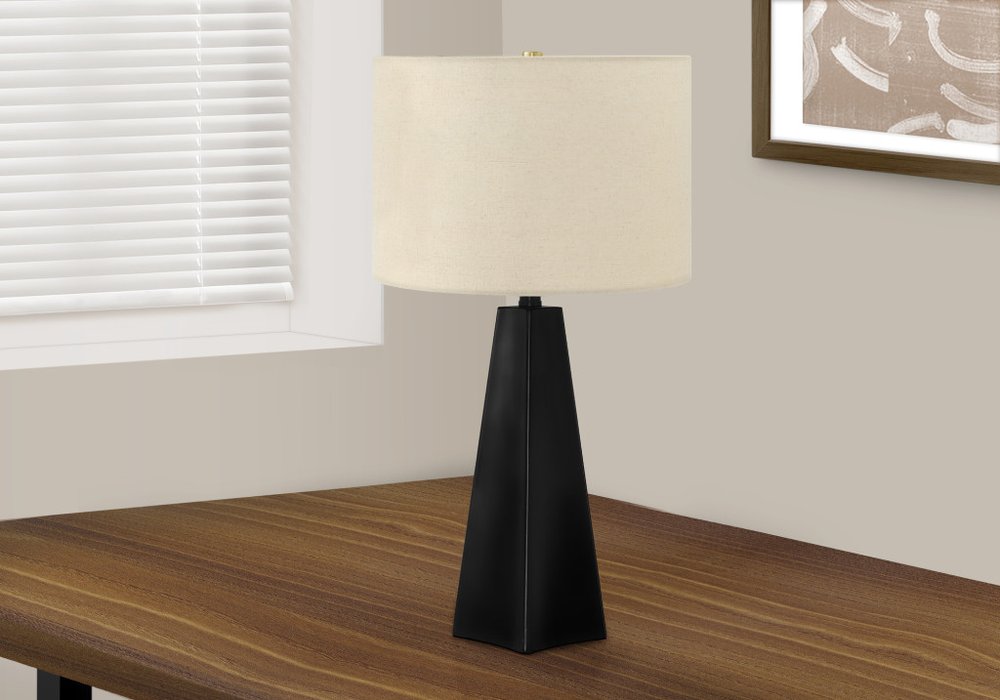 27" Black Geometric Table Lamp With Beige Drum Shade displayed on a dark wood tabletop in a furnished interior against an off-white wall.