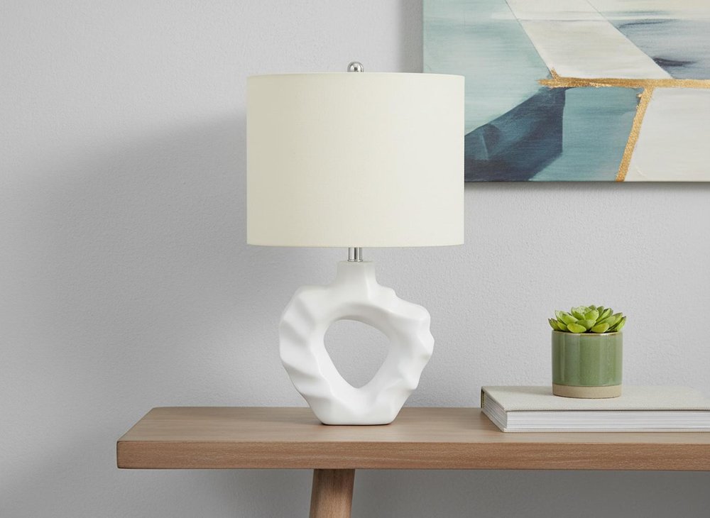 25" Cream Novelty Table Lamp With Cream Drum Shade displayed on a tabletop in a furnished interior against a light gray wall.