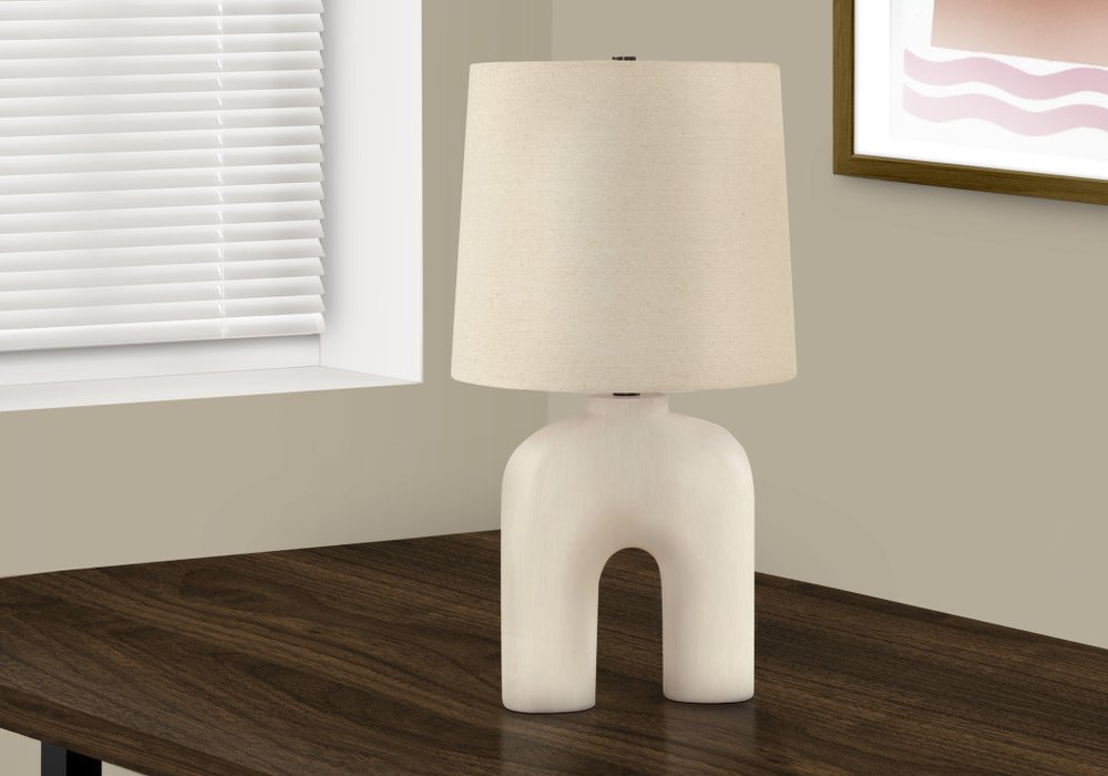 25" Cream Geometric Table Lamp With Beige Empire Shade displayed on a dark wood tabletop in a furnished interior against an off-white wall.