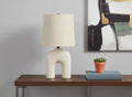 25" Cream Geometric Table Lamp With Beige Empire Shade displayed on a tabletop in a furnished interior against a light gray wall.