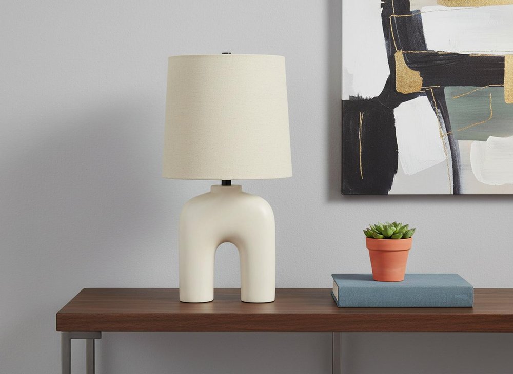 25" Cream Geometric Table Lamp With Beige Empire Shade displayed on a tabletop in a furnished interior against a light gray wall.