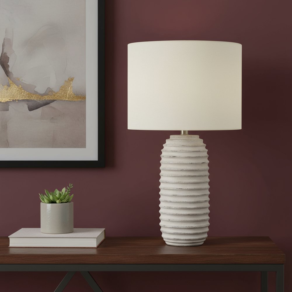 28" Cream Geometric Table Lamp With Cream Drum Shade displayed on a tabletop in a furnished interior against a burgundy wall.