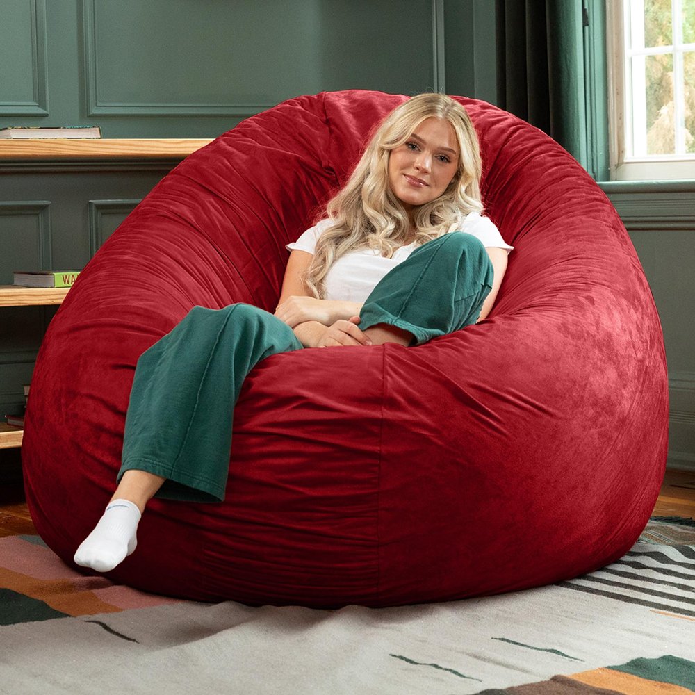 A woman sits in a large, cinnabar red microsuede bean bag chair in a room with a window and a shelf.