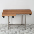 Brown teak wood shower bench with stainless steel legs, mounted on a white tiled wall and grey hexagonal tiled floor.