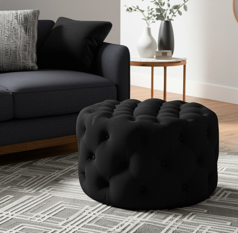 33" Black Linen Tufted Round Cocktail Ottoman styled in a furnished interior with a gray wall on a tiled floor.