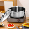 A silver stainless steel stock pot with lid sits on a gray induction cooktop on a wooden table, with food in bowls in the foreground.