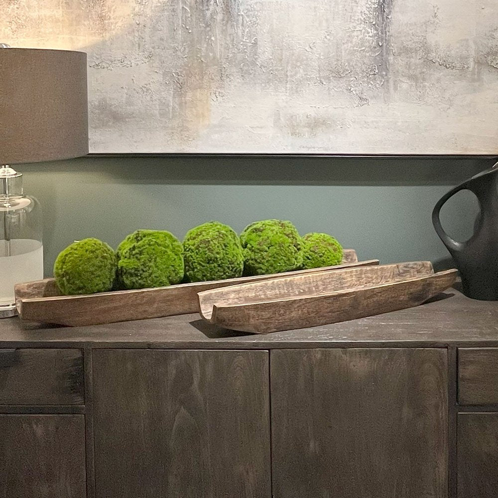 Two dark brown solid wood boat vanity trays with moss balls on a dark wood dresser with a painting and lamp in the background.