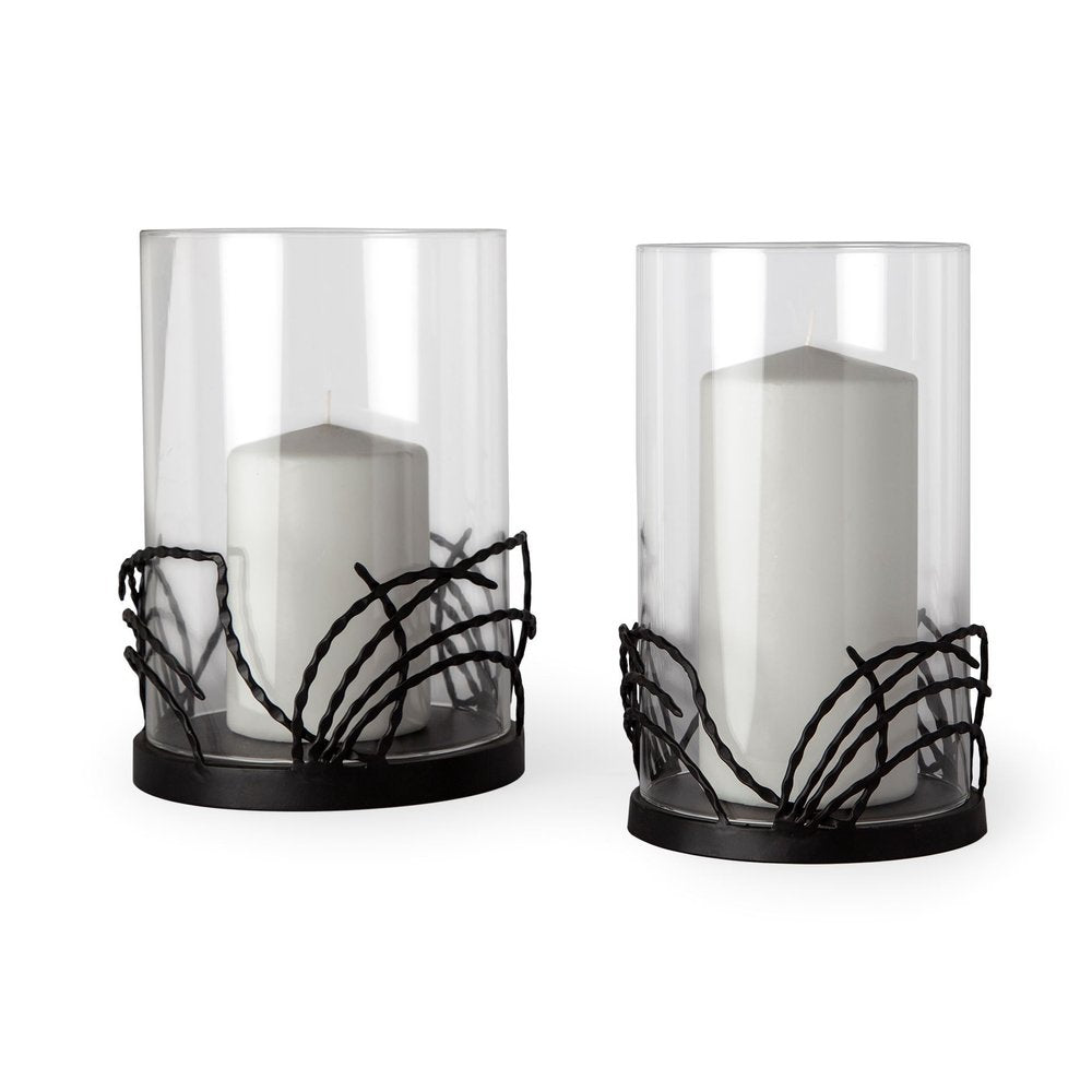 Two clear glass hurricane candle holders with black metal botanical leaf bases and white pillar candles on a white background.