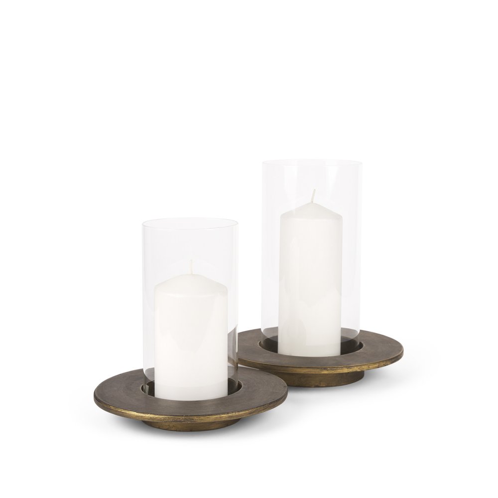 Two white pillar candles in clear glass hurricanes with distressed gold metal bases on a white background.