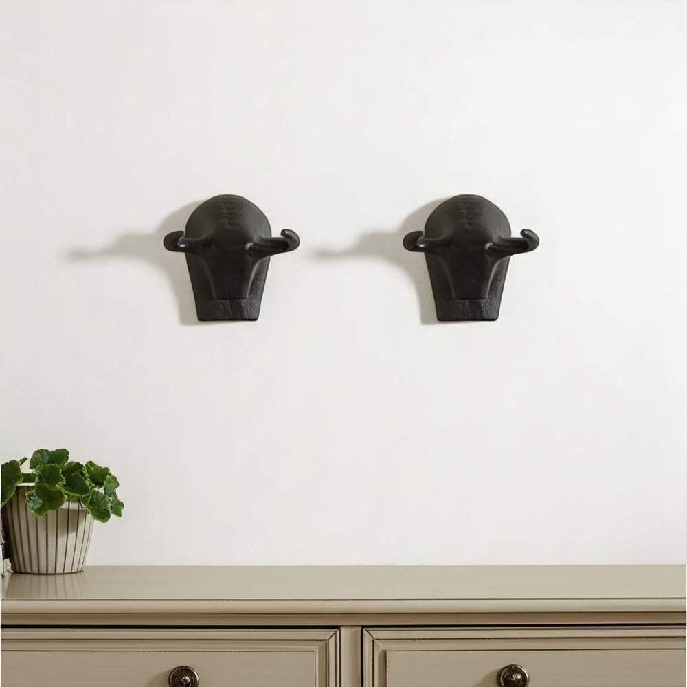Two black metal bull head wall sculptures hang on a white wall above a light gray dresser with a potted plant.