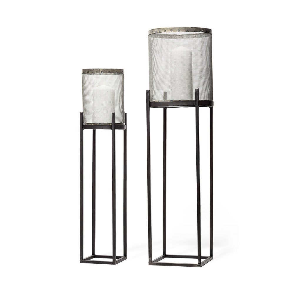 Two black distressed geometric metal lantern candle holders with mesh cylinders and white candles on white background.