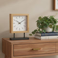 11" Natural And Black Solid Wood And Metal Square Table Clock styled in a furnished interior with a tan wall on a dark wood floor in tan wood.