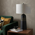 30" Black Round Bedside Lamp With Drum Shade styled in a furnished interior with a tan wall on a dark wood floor.