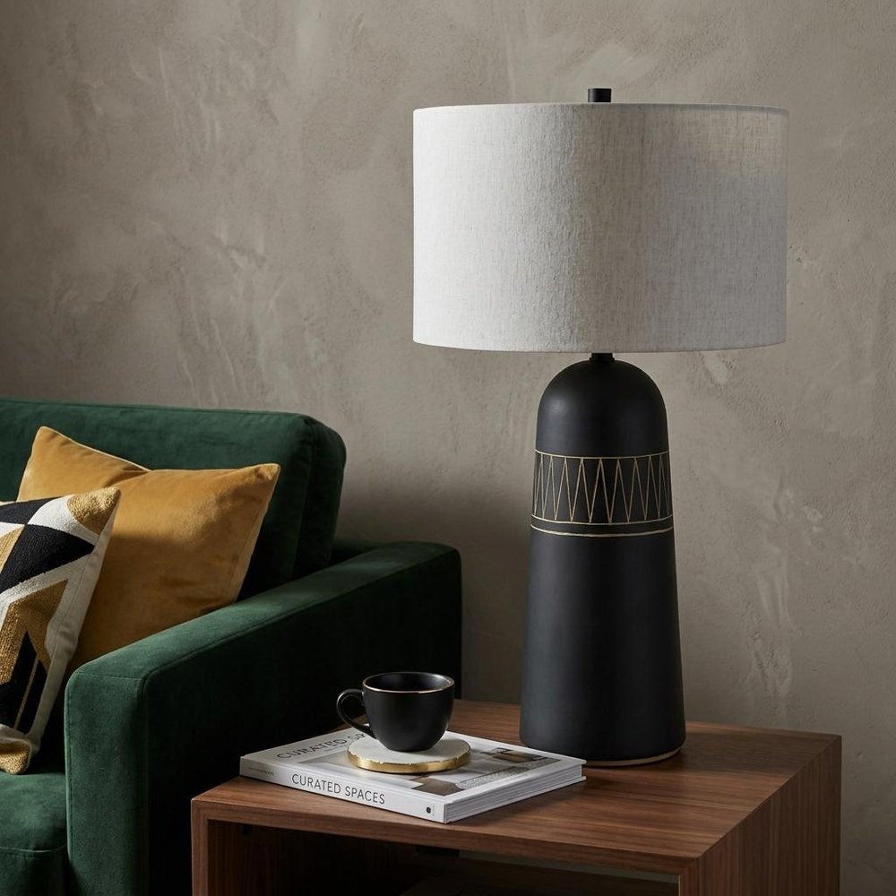30" Black Round Bedside Lamp With Drum Shade styled in a furnished interior with a tan wall on a dark wood floor.