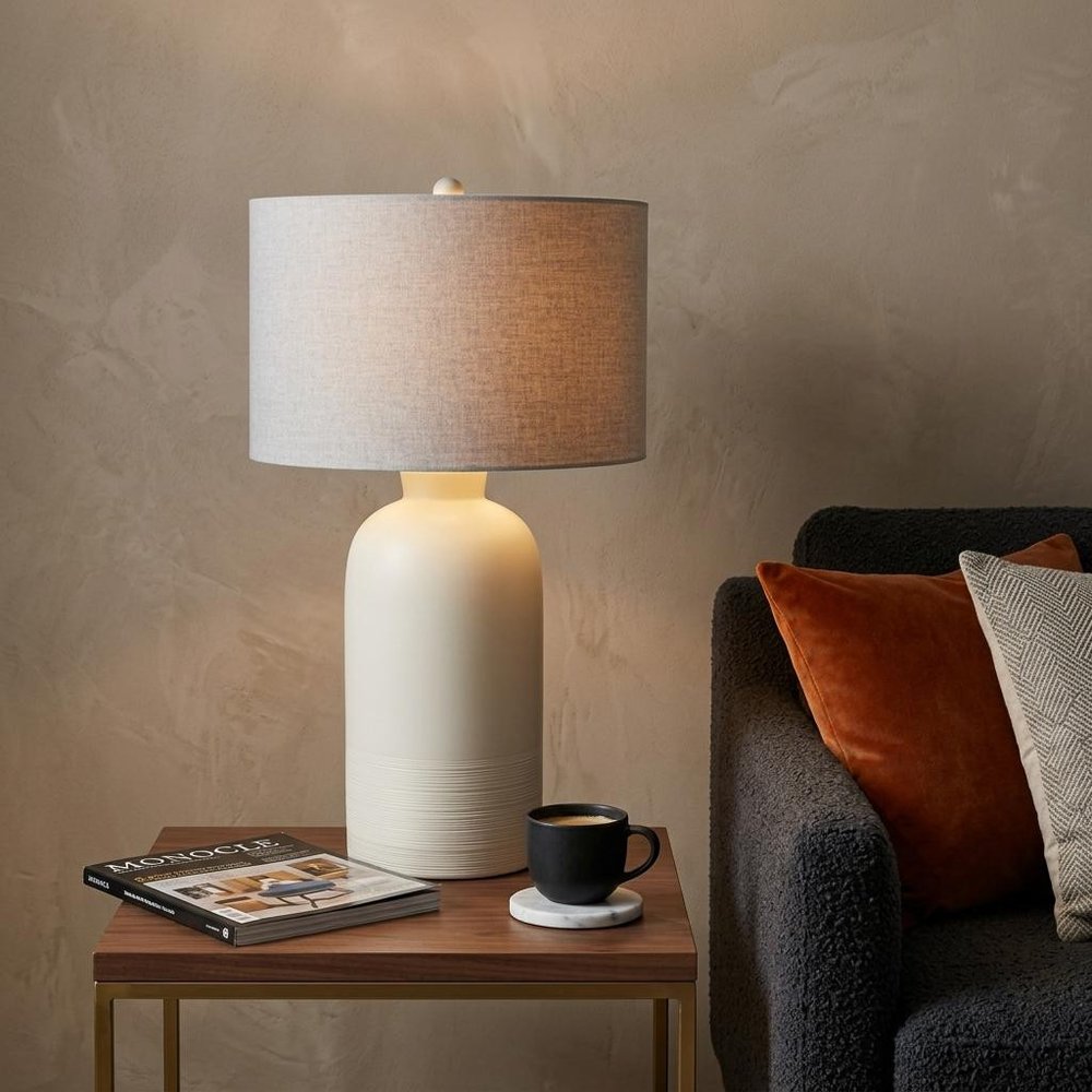 33" Cream Round Bedside Lamp With Drum Shade styled in a furnished interior with a tan wall on a dark wood floor.