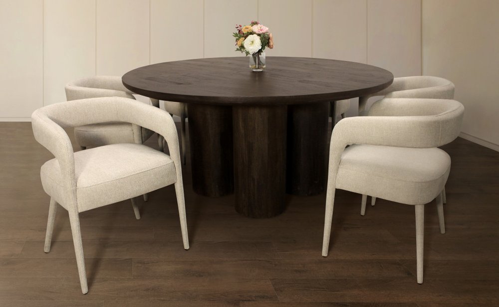 Round dark brown solid wood dining table with six cream upholstered chairs in a room with light walls and dark wood floor.