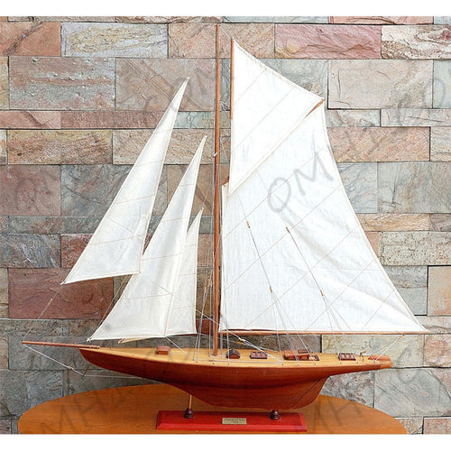 Wooden model sailboat with white sails and a red hull on a wooden stand against a stone wall.