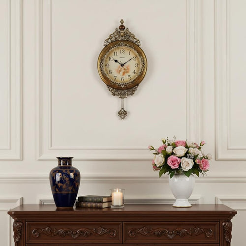 Ornate gold and silver resin round wall clock with pendulum, featuring a floral dial, hanging on a white paneled wall above a dark wood dresser with a blue vase and flowers.