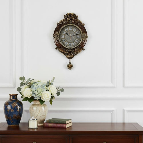 Ornate oval wall clock in brown and silver resin with a pendulum, hanging on a white wall above a wooden dresser with vases and books.