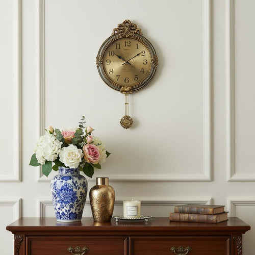 Ornate gold and silver resin round wall clock with pendulum hanging on a white wall above a wooden dresser with flowers and books.