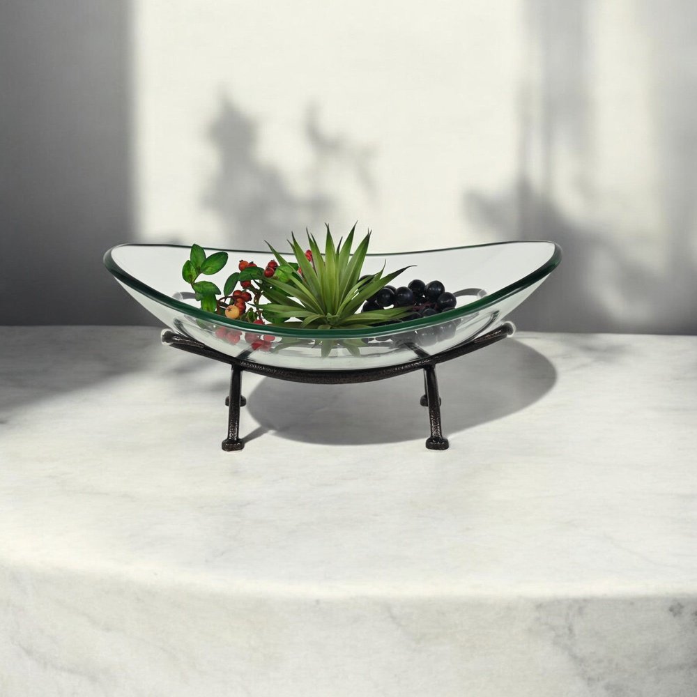 17" Clear And Black Glass And Metal Oval Decorative Bowl With Stand styled in a furnished interior with an off-white wall on a off-white floor.