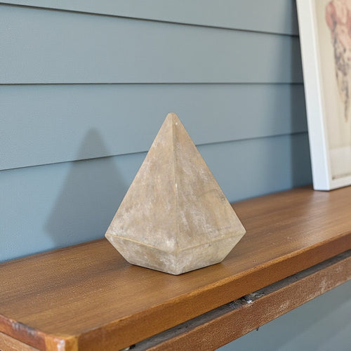 A brown solid wood pyramid decorative sculpture sits on a wooden shelf against a blue paneled wall.