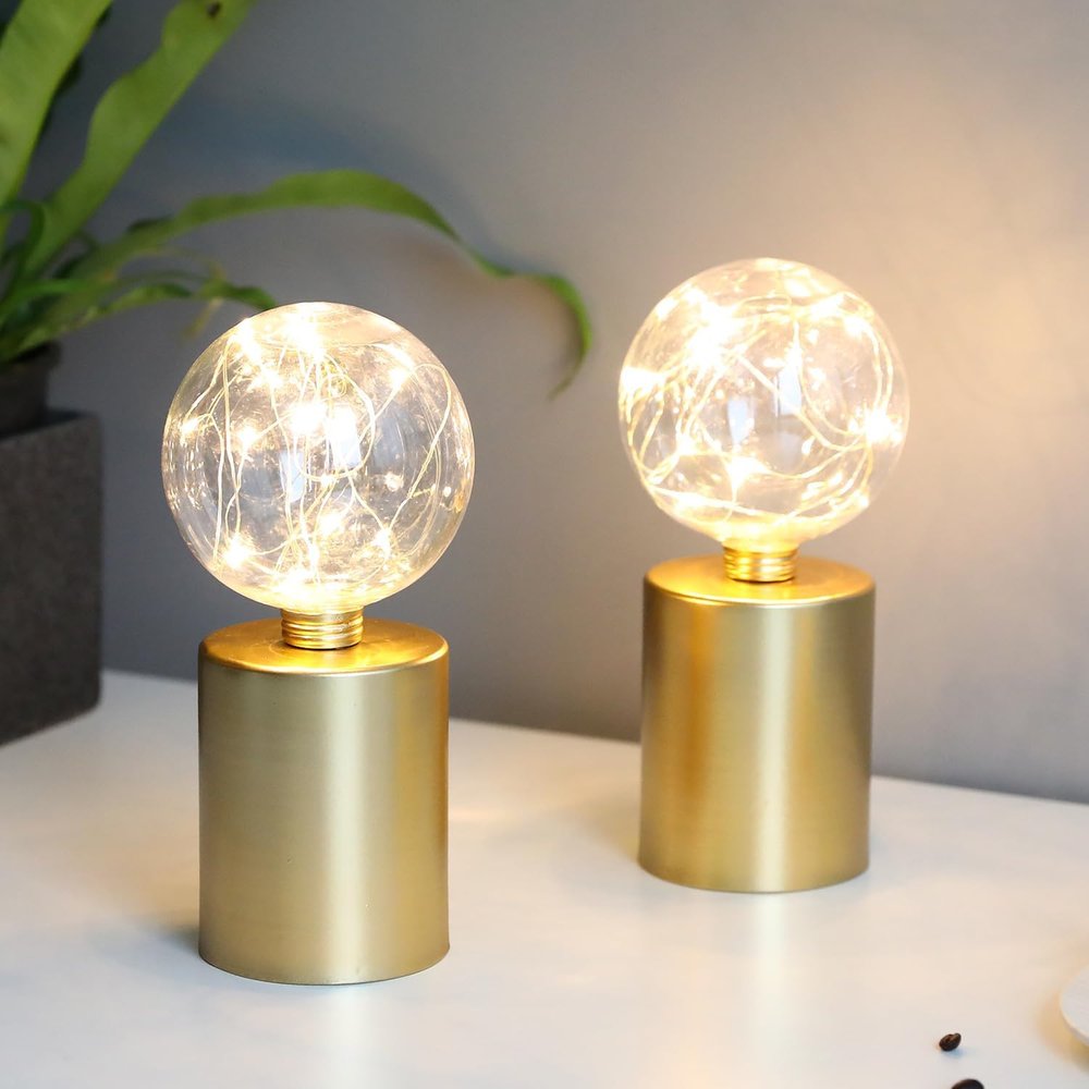 Two gold bedside lamps with clear globe shades containing fairy lights on a white surface with a plant in the background.