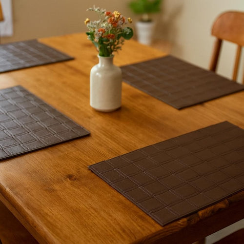 Espresso basketweave faux leather placemats on a wooden table with a vase of flowers.