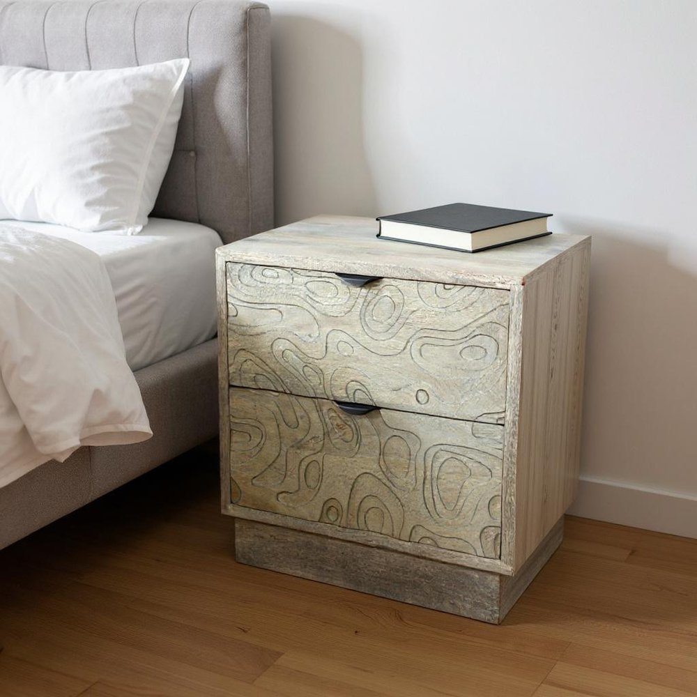 26" Gray Solid Wood Carved Abstract Two Drawer Nightstand styled in a furnished interior with an off-white wall on a dark wood floor.