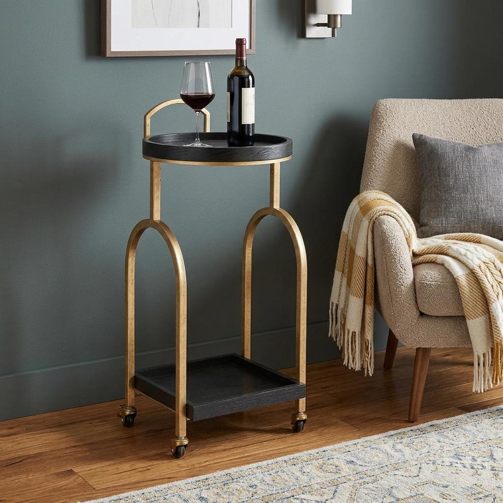 17" Black And Brass Steel and Solid Wood Distressed Round Rolling Bar Cart styled in a furnished interior with a gray wall on the floor.