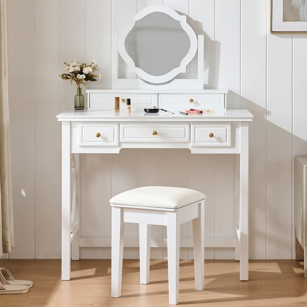 White wood vanity table with a scalloped mirror and stool, featuring five drawers and makeup accessories on a wood floor.