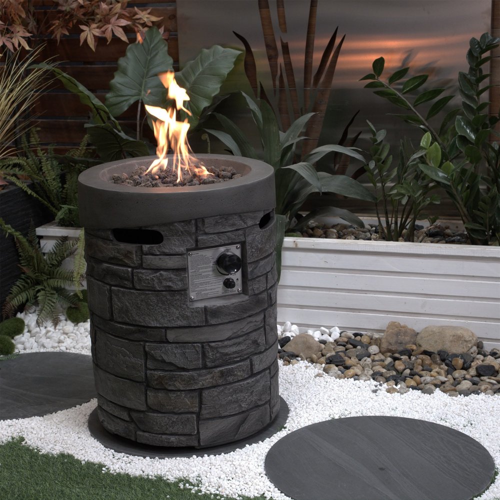32000 BTU 26" Gray Magnesium Oxide Propane Round Fire Pit With Cover styled in a furnished interior with a dark wall on a charcoal floor.