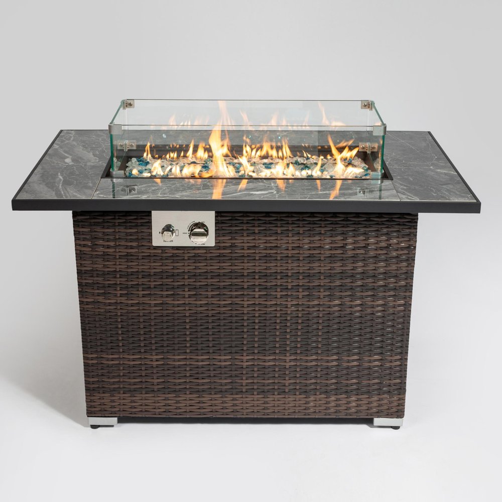 Rectangular propane fire pit with brown wicker base, gray stone top, and glass wind guard with flames.