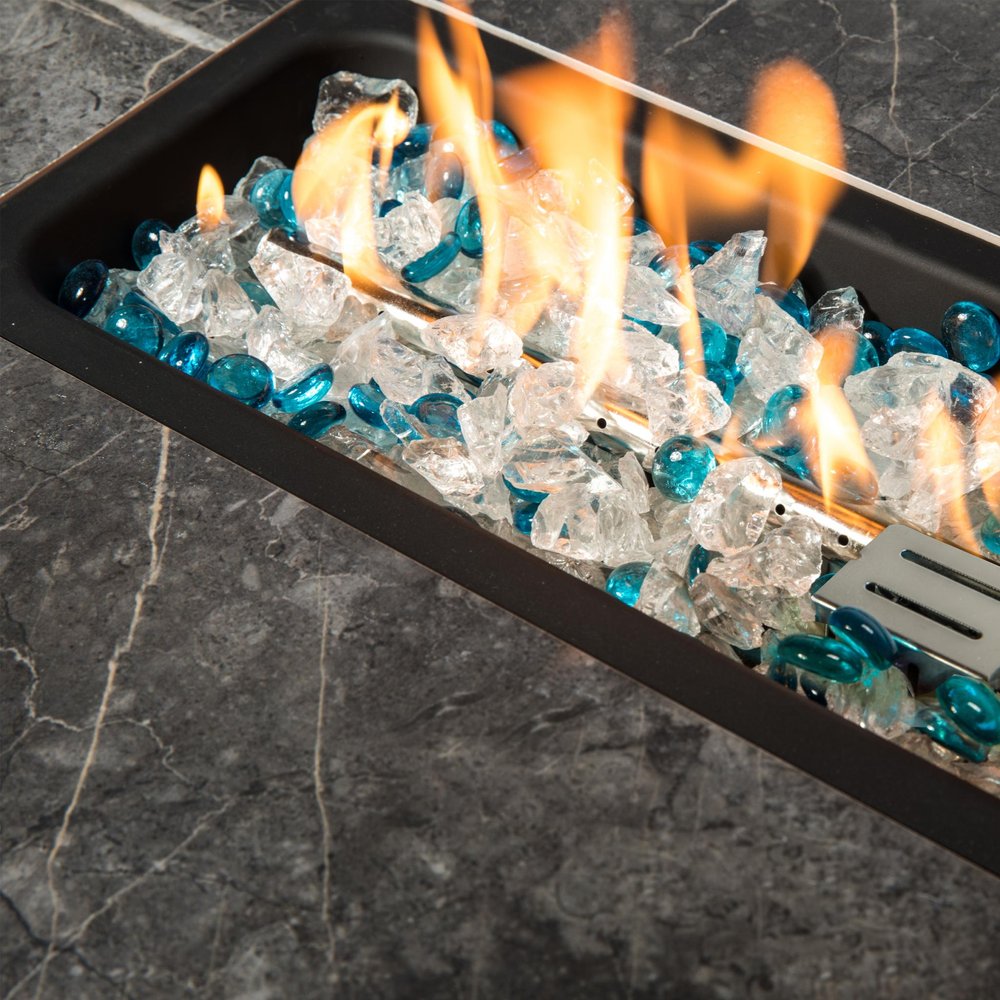 Close-up of a rectangular fire pit with clear and blue glass rocks and flames on a dark marble-like surface.