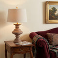 27" Brown Urn Bedside Lamp With Drum Shade styled in a furnished interior with a cream wall on a black floor.