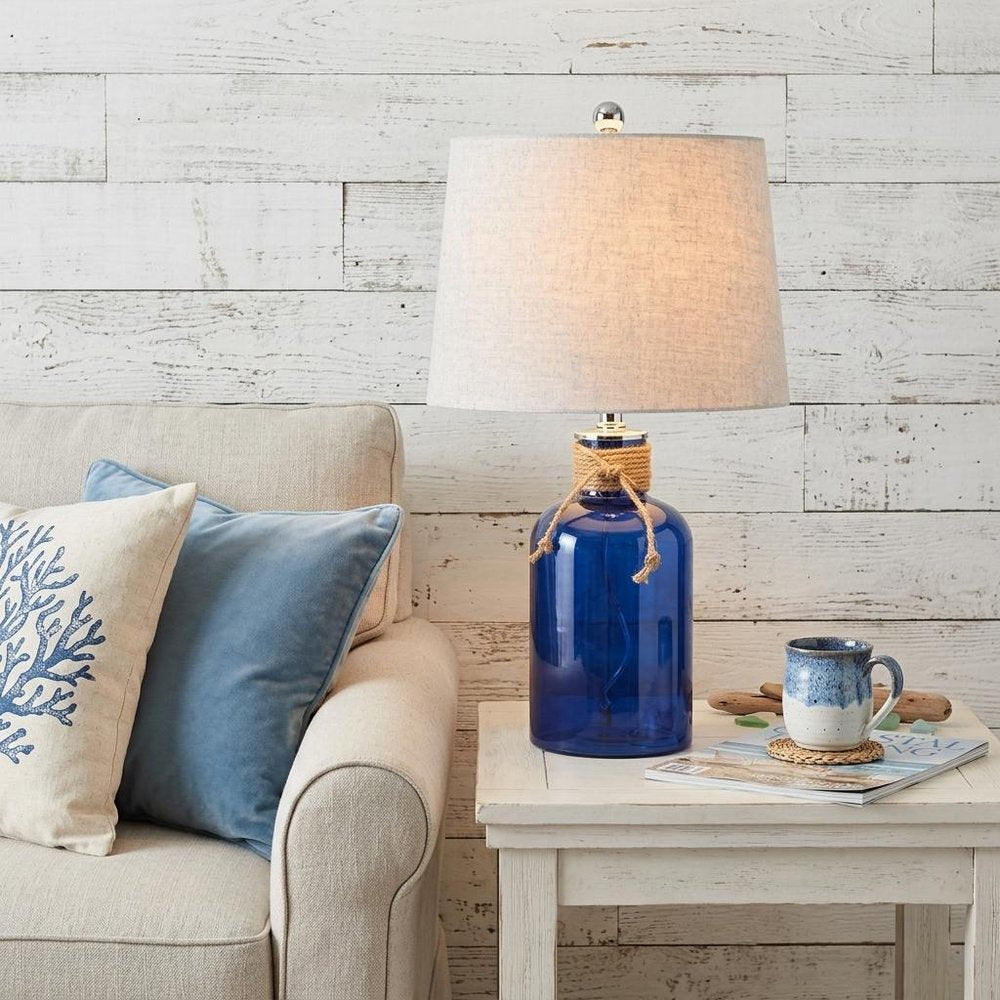 24" Clear Novelty Table Lamp With Drum Shade displayed on a wood tabletop in a furnished interior against an off-white wall.