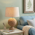 24" Brown Globe Table Lamp With Drum Shade displayed on a wood tabletop in a furnished interior against a cream wall.